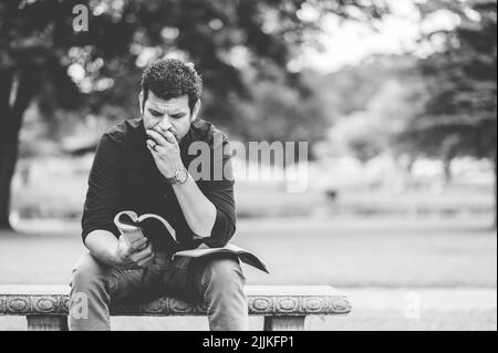 A closeup of a man reading a book and sitting on a bench in a park Stock Photo