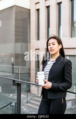 Confident young Asian female manager in formal clothes with reusable tumbler for takeaway drink leaning on glass fence among office buildings Stock Photo