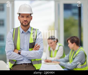 I trust my team completely. Cropped portrait of a handsome young male construction worker standing with his arms folded with his colleagues in the Stock Photo