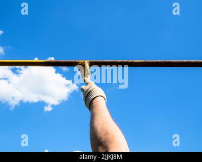 hand paints rusty outdoor gas pipe with yellow paint using brush and blue sky on background on sunny summer day Stock Photo
