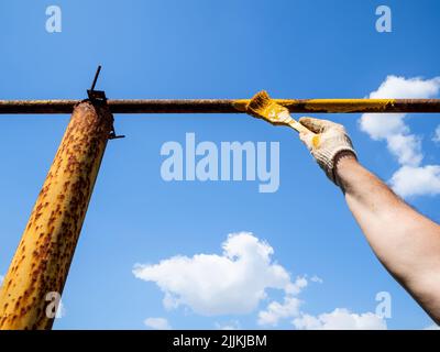 hand paints rusty outdoor gas pipeline with brush and blue sky on background on sunny summer day Stock Photo