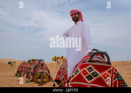 An Arab male in a twabh standing near his camels on a desert Stock Photo