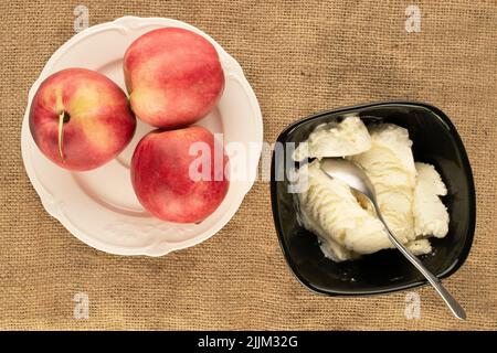 Several balls of white ice cream in a black ceramic dish and three nectarines with a white plate on jute fabric, macro, top view. Stock Photo