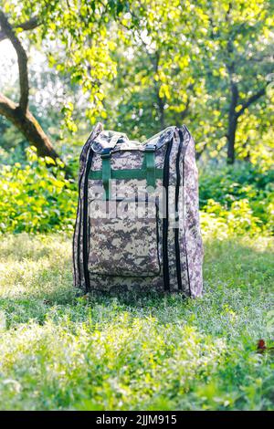 Defocus military backpack. Army bag on green grass background near tree. Military camouflage webbing material on a British army rucksack. Vertical. To Stock Photo