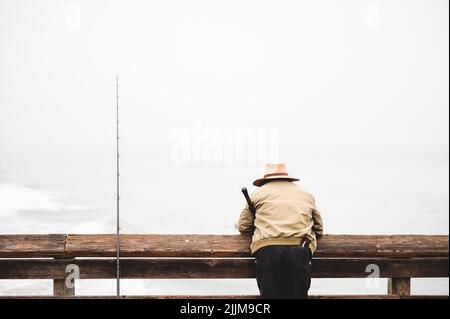 A rear view of a fisherman leaning on a wooden railing of a dock looking at the coast Stock Photo