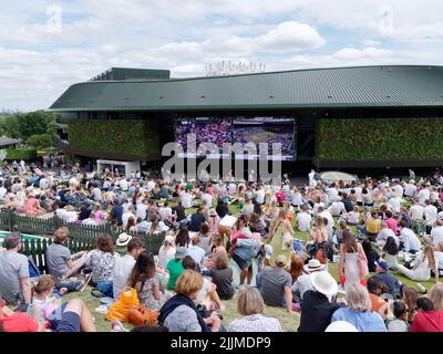 Wimbledon Tennis Championship. Crowds sit on the lawn on The Hill outside number one court watching the action on a big screen. London.