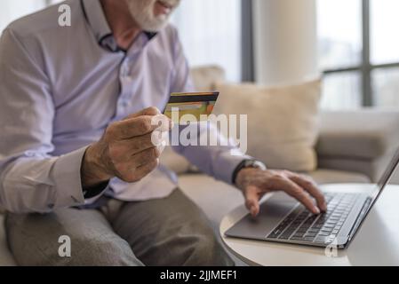 Midsection of senior business professional making online payment through credit card. Male entrepreneur is using laptop at table. He is sitting in hom Stock Photo