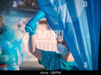 April 6 2019 Tulsa USA - Holi - Color Run in Tulsa USA - Volunteer in mask  standing behind flag squirts turquoise powder on runner against blurred ba Stock Photo