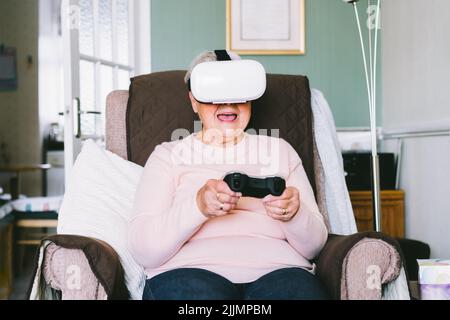 Emotional senior woman wearing virtual reality goggles and using wireless controller, joystick gamepad playing video games at home. Fun on Retirement. Stock Photo