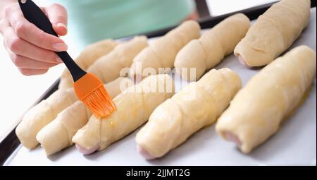 Female cook greases sheet of dough with melted butter and prepares sausage wrapped in dough Stock Photo