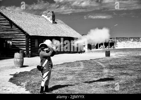 A grayscale shot of a man wearing clothes of a french soldier and shooting from the gun. Fort Niagara. Stock Photo