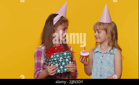 Happy little toddler children sister girls siblings friends wearing festive cone cap celebrating birthday anniversary party, blowing candle on small cake cupcake making a wish, dancing. Preschool kids Stock Photo
