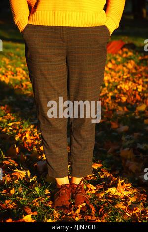 Defocus female legs in capri pants and brogues shoes on green grass with bright leaves. Bright stylish woman in orange coat walking in october park. O Stock Photo