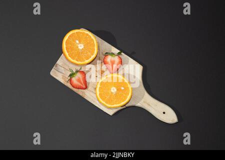 A top view of a wooden board with a halved orange and a halved strawberry on a black surface Stock Photo