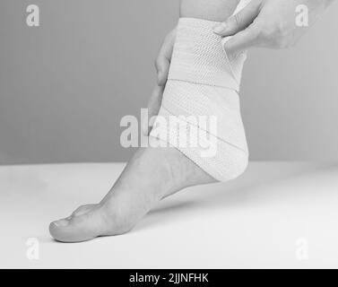 Woman wrapping elastic bandage around painful ankle to relieve pain or prevent injury. Feet trauma. Sprains, strains, tendonitis treatment. Health problems, medical conditions. Black and white. photo Stock Photo