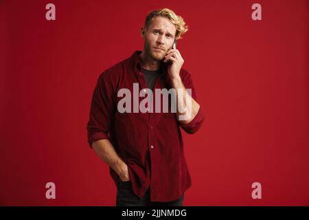 White bearded man frowning while talking on mobile phone isolated over red wall Stock Photo