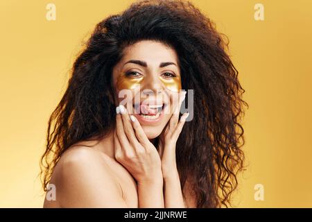 Enjoyed happy pretty Latin female with gold hydrogel patches under eye, stick tongue touch cheek posing isolated over yellow background. Cosmetic Stock Photo
