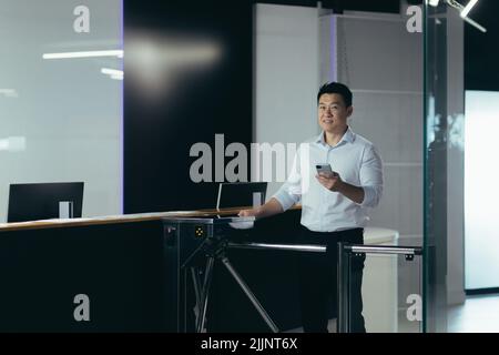 A young handsome Asian businessman, an office worker goes to the office and the business center for work in the morning through the security counter and automatic electronic pass. He holds the phone Stock Photo
