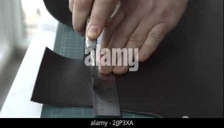 Working process of crafting a new handmade leather wallet in the leather workshop. Man's hands working with knife and iron ruler and cutting a leather Stock Photo