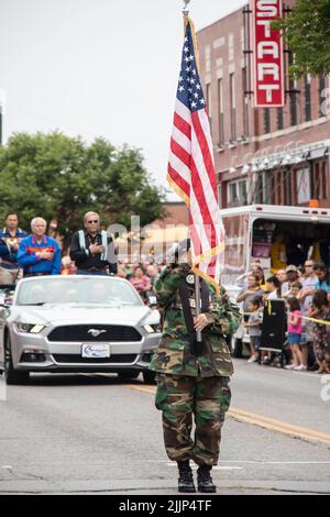 Tahlequah USA 8 31 2019 Member of Cherokee Honor Guard in camos presents American Flag in parade while tribe elders in Mustang convertible and crowd s Stock Photo