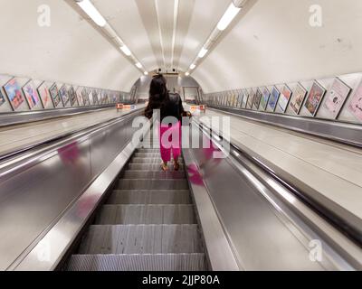 London, Greater London, England, July 02 2022: Lady with pink trousers and black jacket on an escalator on an otherwise empty tube station Stock Photo