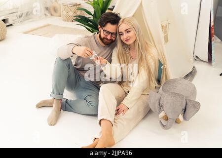 Young expecting white couple looking affectionately at knitted baby shoe sitting on the floor next to tipi tent and elephant plushie. Indoor shot. High quality photo Stock Photo