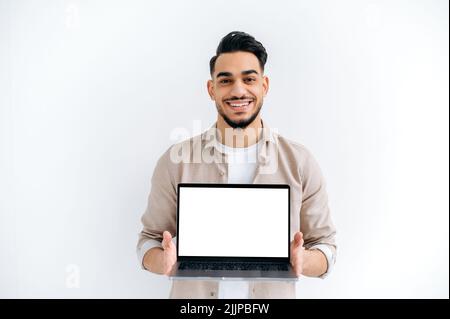 Joyful handsome Indian or Arabian young man, student or freelancer, stands on white isolated background, holding open laptop with blank white mockup screen for presentation, looking at camera, smiling Stock Photo