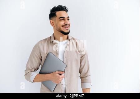 Cheerful arabian or indian young man with beard, wearing casual shirt, standing over isolated white background, holding laptop, looking to the side, smiling joyfully, dreaming, thinking Stock Photo