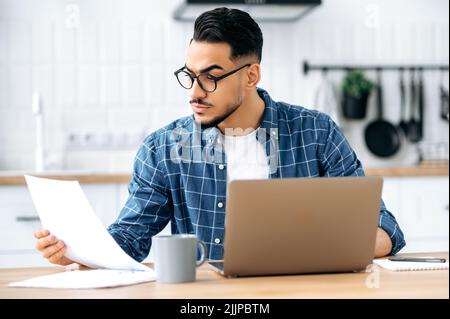 Focused smart successful Indian or Arabian guy, stylishly dressed, with glasses, freelancer, company ceo, working remotely from home in a laptop, sitting in the kitchen, studying documents, reports Stock Photo