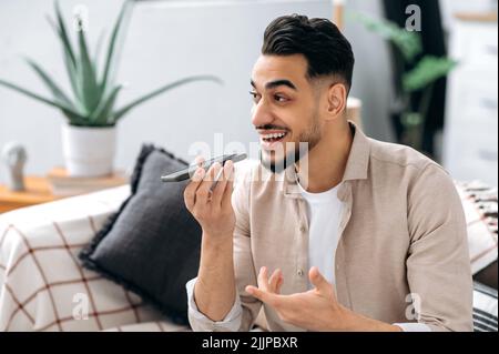 Excited positive indian or arabian man, sitting on a sofa in living room, holding his smartphone, talking on speakerphone and smiling, record voice message. Wireless technology concept Stock Photo