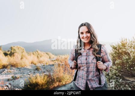 Smiling happy latin young woman with a backpack on, hiking in a beautiful landscape. Stock Photo
