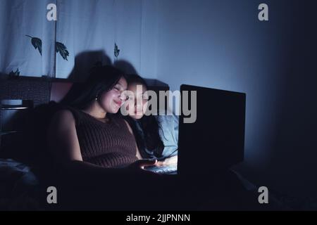 A vertical shot of two Hispanic Young women laying on the bed, watching movies at the dark on a laptop Stock Photo