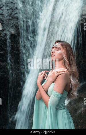 A vertical shot of a young female in a green dress and forehead jewelry posing near a waterfall Stock Photo