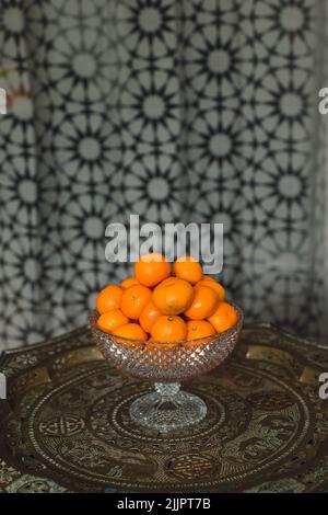 pile of tangerines, satsumas, mandarins oranges in crystal hobnail bowl on brass table with middle eastern star pattern curtain background Stock Photo