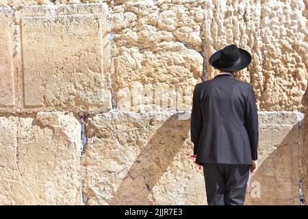 A man standing in a suit and a hat looking at a rock wall in the sun Stock Photo