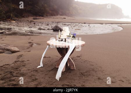 The wedding cake with flowers and decorations on a wooden table on a sandy beach Stock Photo