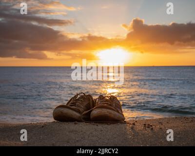 A pair of sneakers on a sandy beach with the Sun reflecting on the ocean at sunset Stock Photo