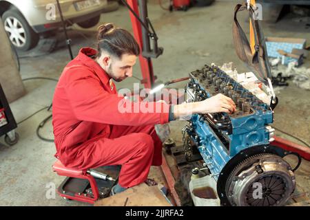 A young Caucasian mechanic in a red uniform mounting the car engine Stock Photo