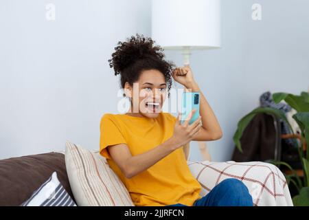 Excited young African American woman using mobile phone shopping online sitting at home on the couch Stock Photo