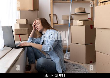 Young worried unhappy woman sitting on the floor near moving boxes using a laptop with hands on head suffering of headache, searching for a new Stock Photo