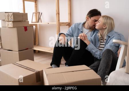 Happy young couple sitting on the floor of the newly rented or purchased apartment. Unpacked cardboard boxes and covered furniture in the modern Stock Photo