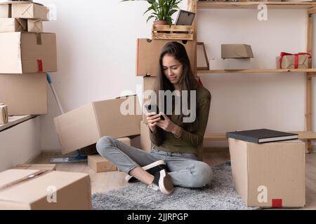 Happy young Hispanic woman using smartphone in living room at new house with stack of cardboard boxes on moving day Stock Photo
