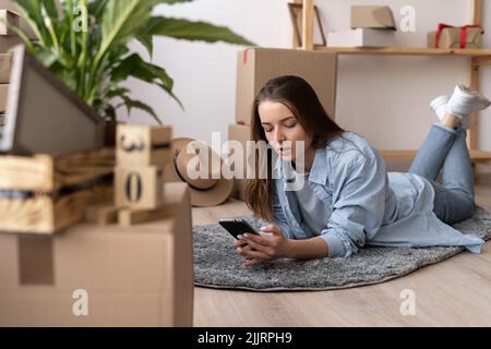 young caucasian woman using smartphone in living room at new house with stack of cardboard boxes on moving day Stock Photo
