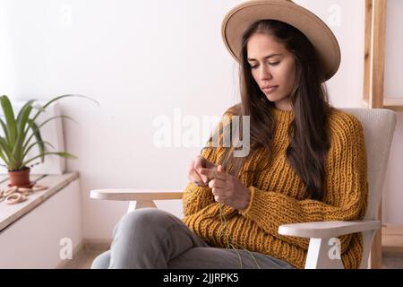 Young girl knits a sweater with knitting needles, homemade needlework from woolen threads, knit clothes at home, female knitter in a hat, hobby and Stock Photo