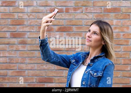 Photo of cheerful cute charming nice beautiful attractive girlfriend blonde, pale skinned taking selfie with her telephone held in her hands wearing jeans jacket isolated over red color brick background. High quality photo Stock Photo