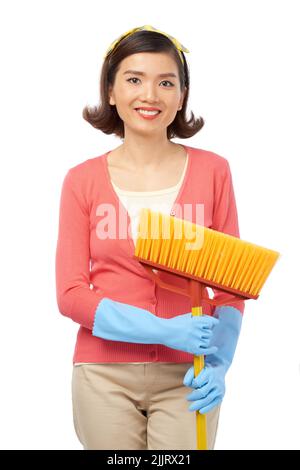 Portrait shot of beautiful Asian woman with broom in hands distracted from sweeping floor in order to pose for photography, isolated on white background Stock Photo