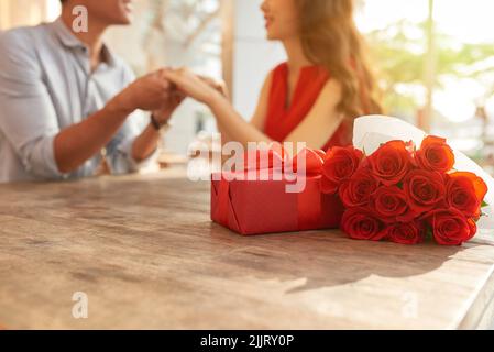 Cheerful young couple sitting at wooden table of outdoor cafe and holding hands while celebrating Valentines Day, gift box and bouquet of red roses on foreground Stock Photo