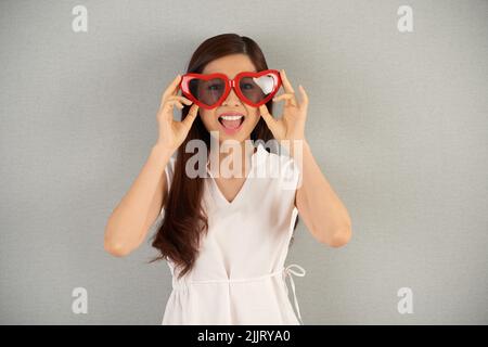 Waist-up portrait of cheerful Asian woman wearing large heart-shaped sunglasses looking at camera while standing against gray background Stock Photo