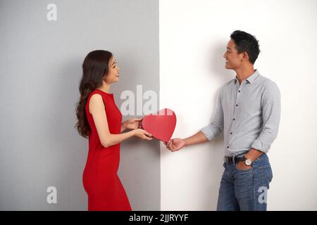 Profile view of joyful Asian boyfriend and girlfriend looking at each other with toothy smiles and holding heart shaped gift box in hands while standing against gray and white background Stock Photo