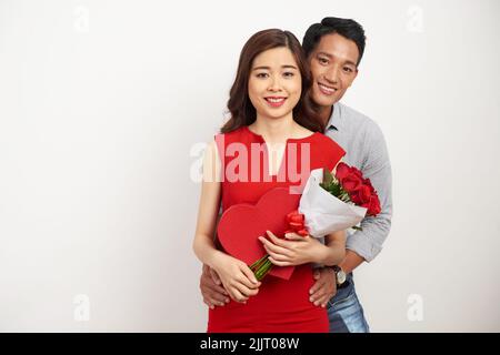 Group portrait of cheerful Asian couple looking at camera with wide smiles while celebrating Valentines Day together, pretty woman holding bouquet of red roses and heart shaped gift box in hands Stock Photo